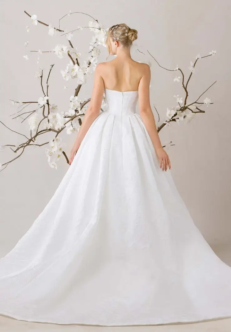 A bride in a strapless white gown stands with her back to the viewer, surrounded by delicate white flowers on branches, exuding elegance and serenity.