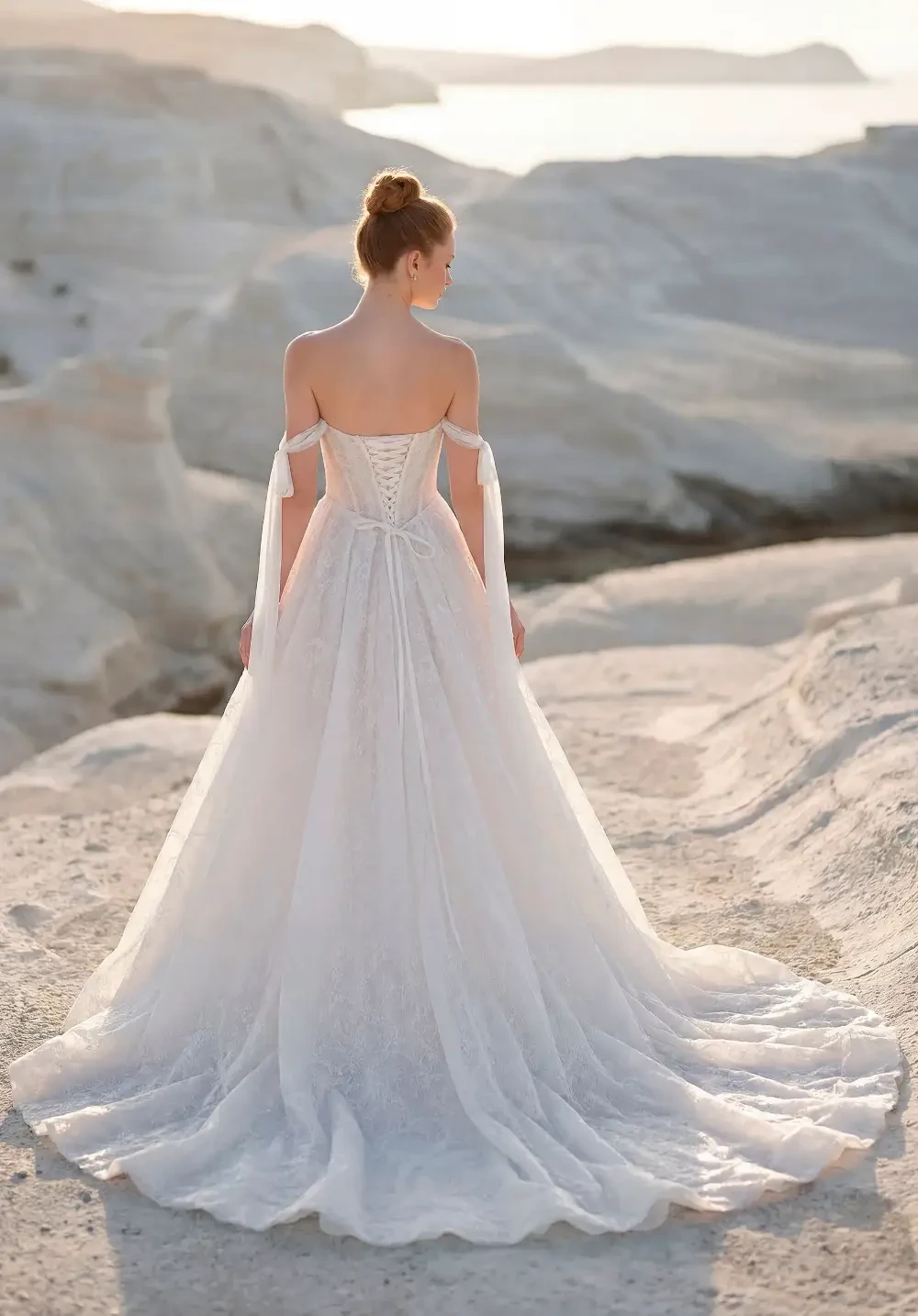 A woman in a flowing white dress stands on a rocky beach with the ocean in the background.