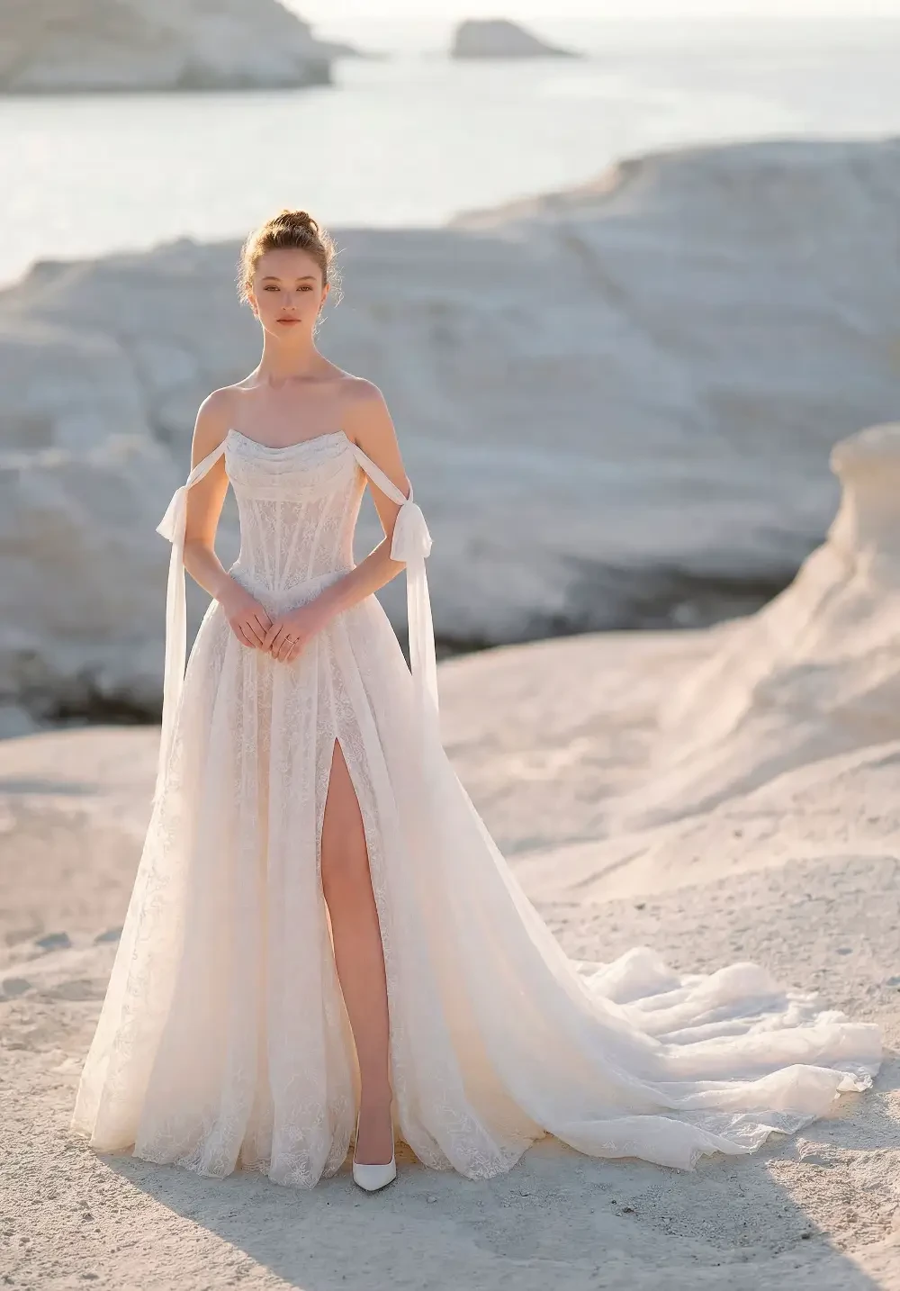 A woman in a flowing white dress stands on a rocky beach with the ocean in the background.
