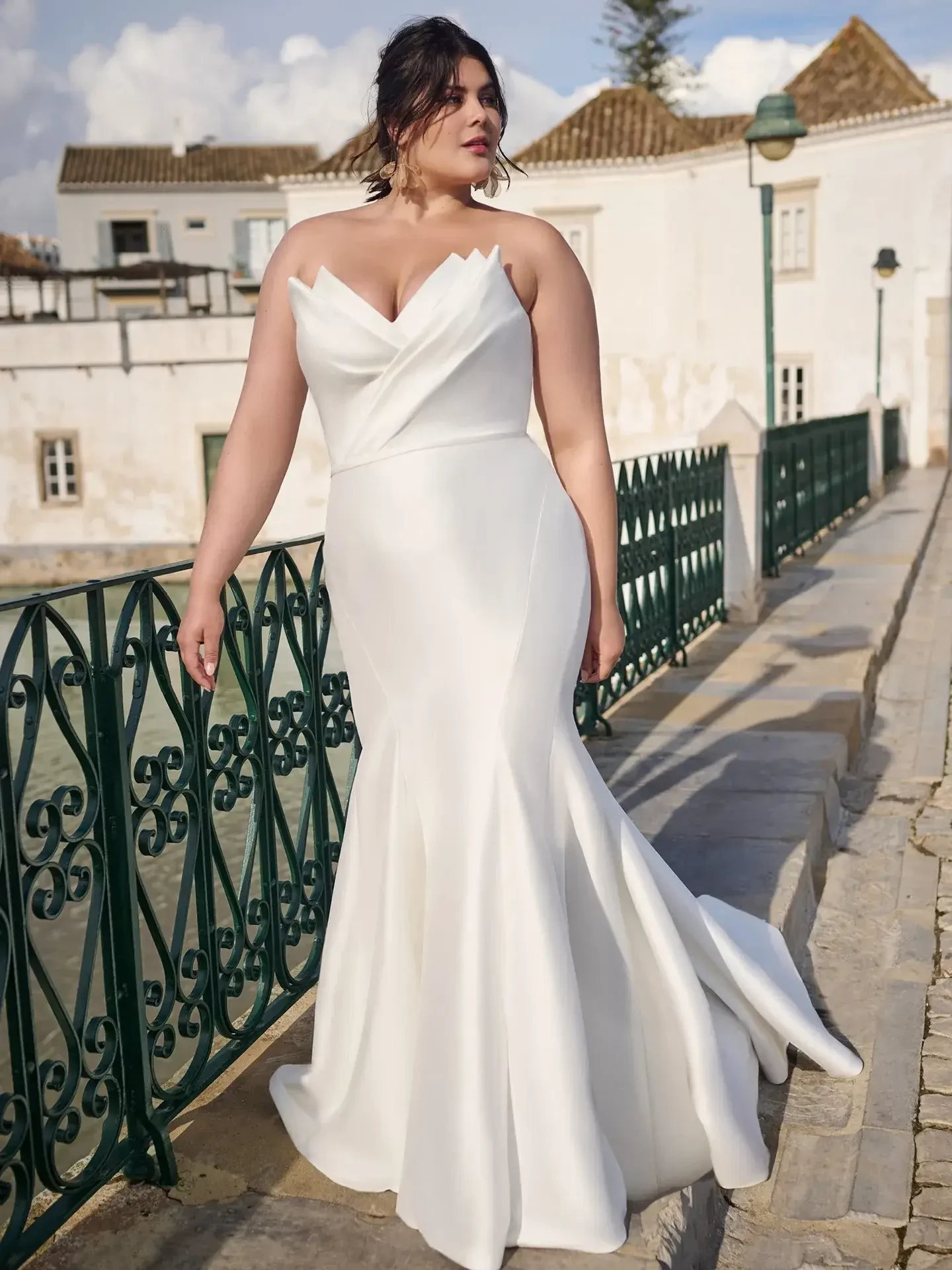 A woman in a form-fitting white wedding dress stands on a cobblestone path near a green railing, with white buildings and trees in the background.