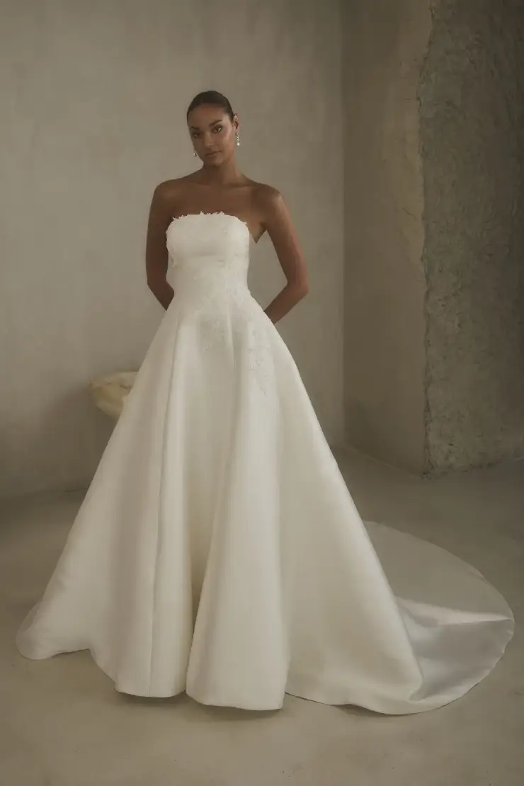A woman in an elegant strapless wedding dress with a full skirt and subtle floral details stands in a minimalist room, conveying timeless grace.