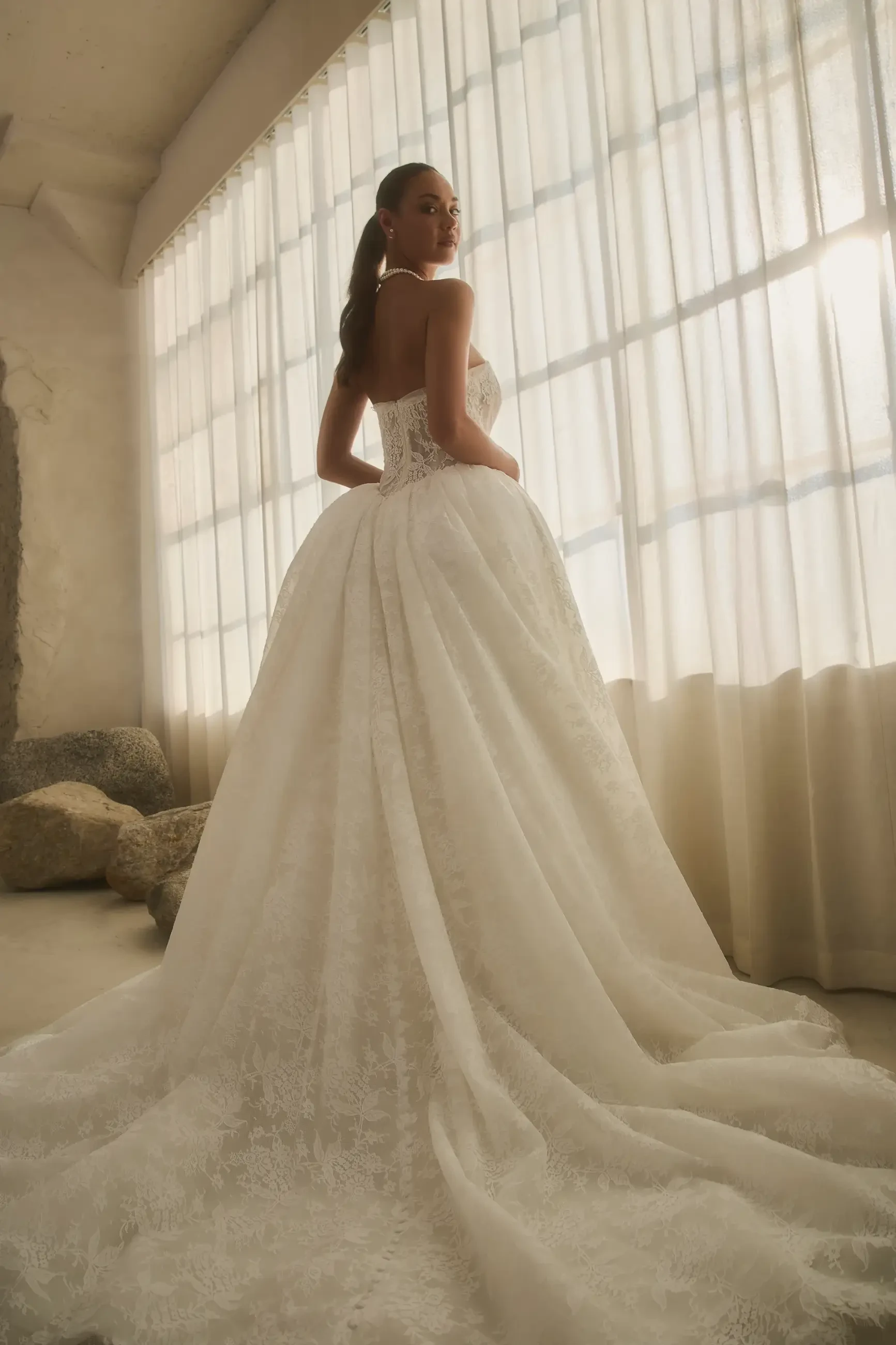 A woman in a strapless, lace wedding gown with a full skirt and sheer paneling stands in front of soft, flowing curtains. She exudes elegance and sophistication.