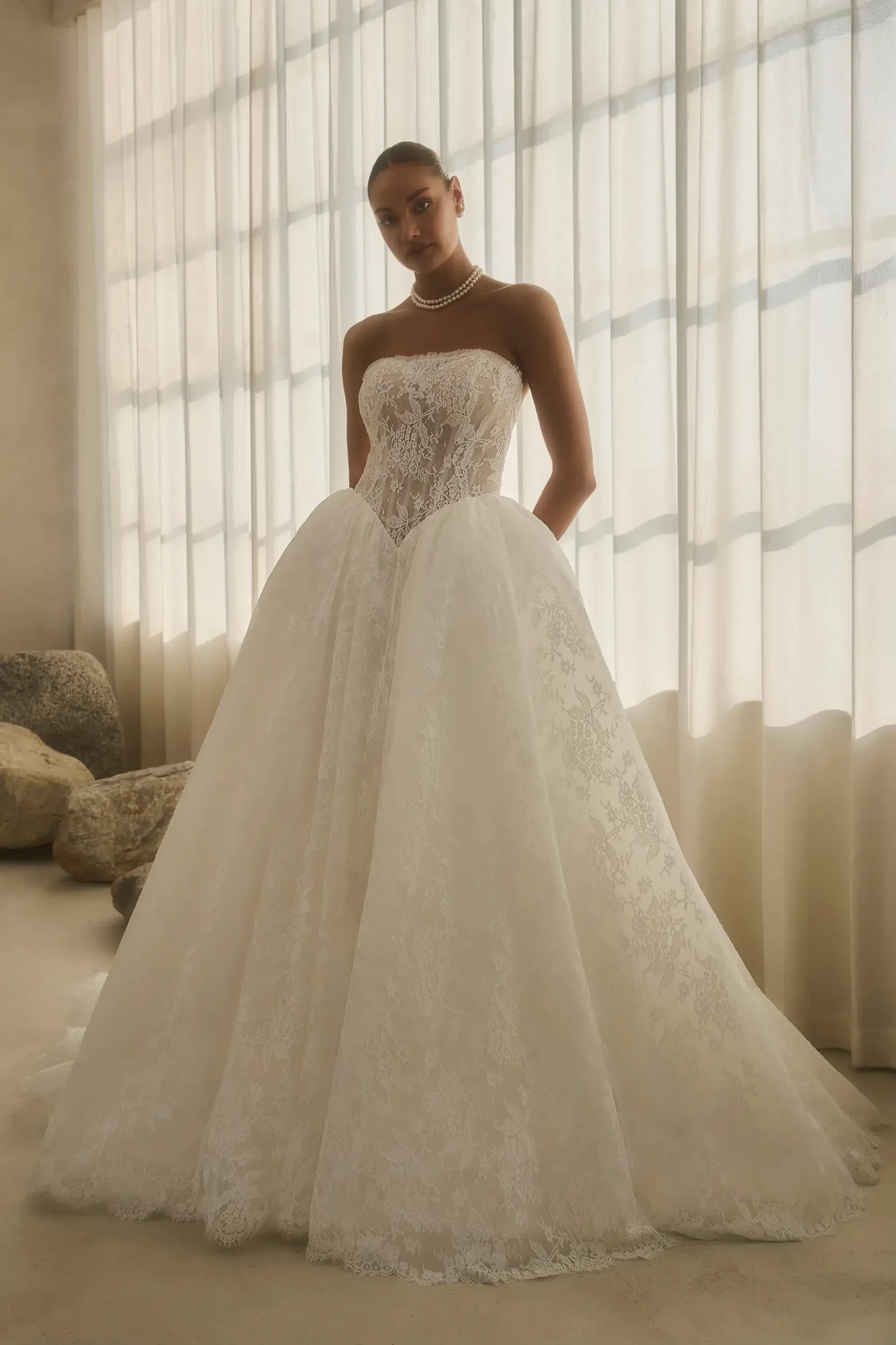 A woman in a strapless, lace wedding gown with a full skirt and sheer paneling stands in front of soft, flowing curtains. She exudes elegance and sophistication.