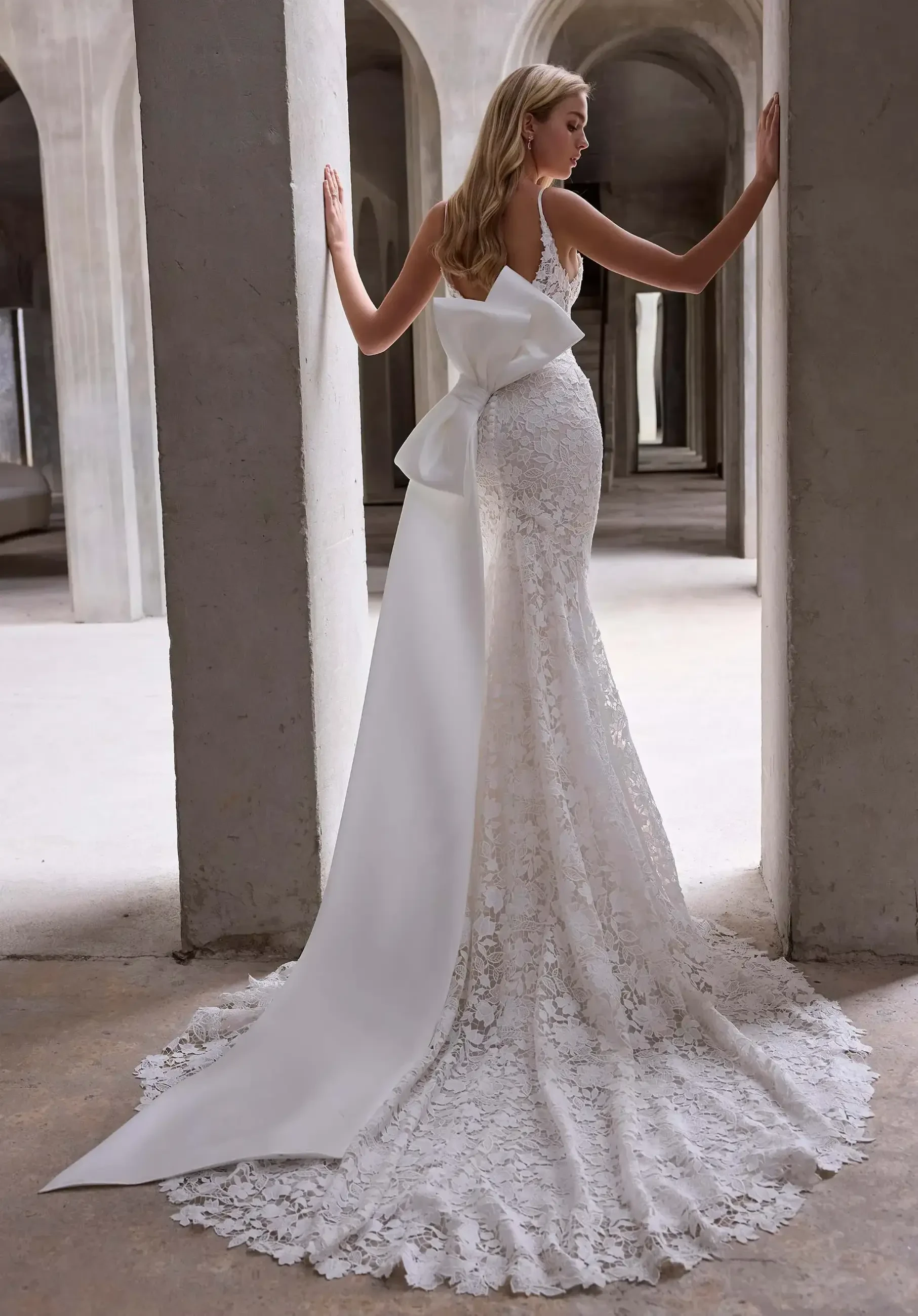 A woman wearing a fitted white lace wedding dress with a long train and a large bow at the back, standing in an architectural space with columns.