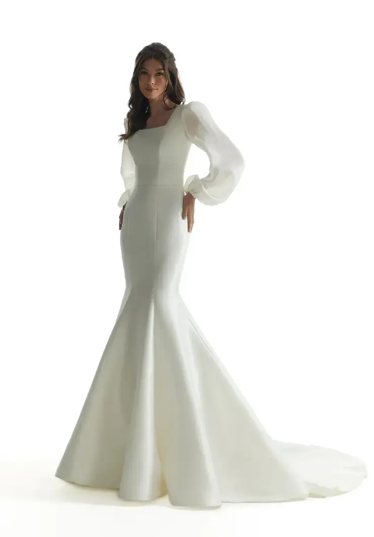 A woman in an elegant white mermaid-style gown with sheer puff sleeves stands against a white background, exuding a graceful and sophisticated aura.