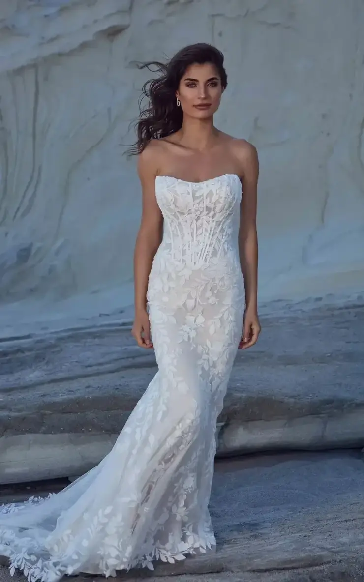 A woman wearing a strapless, fitted white wedding dress with intricate lace details, standing on a rocky surface.