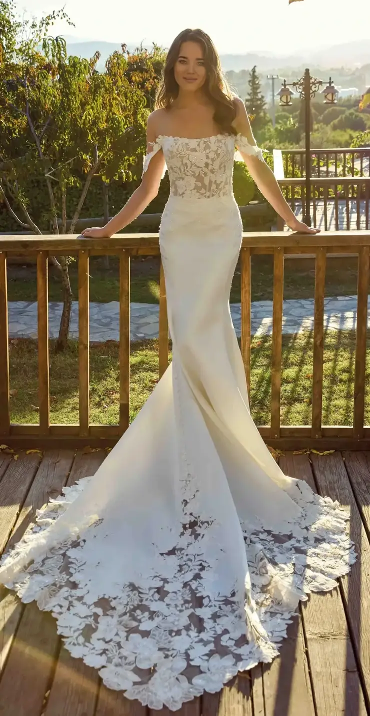 A woman in an elegant white wedding dress with off-the-shoulder sleeves stands on a wooden deck, smiling at the camera. The dress features intricate lace detailing at the bodice and a dramatic train. In the background, there are trees and a scenic view of hills and sunlight.