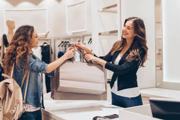 Smiling sales associate handing a shopping bag to a customer in a clothing store.