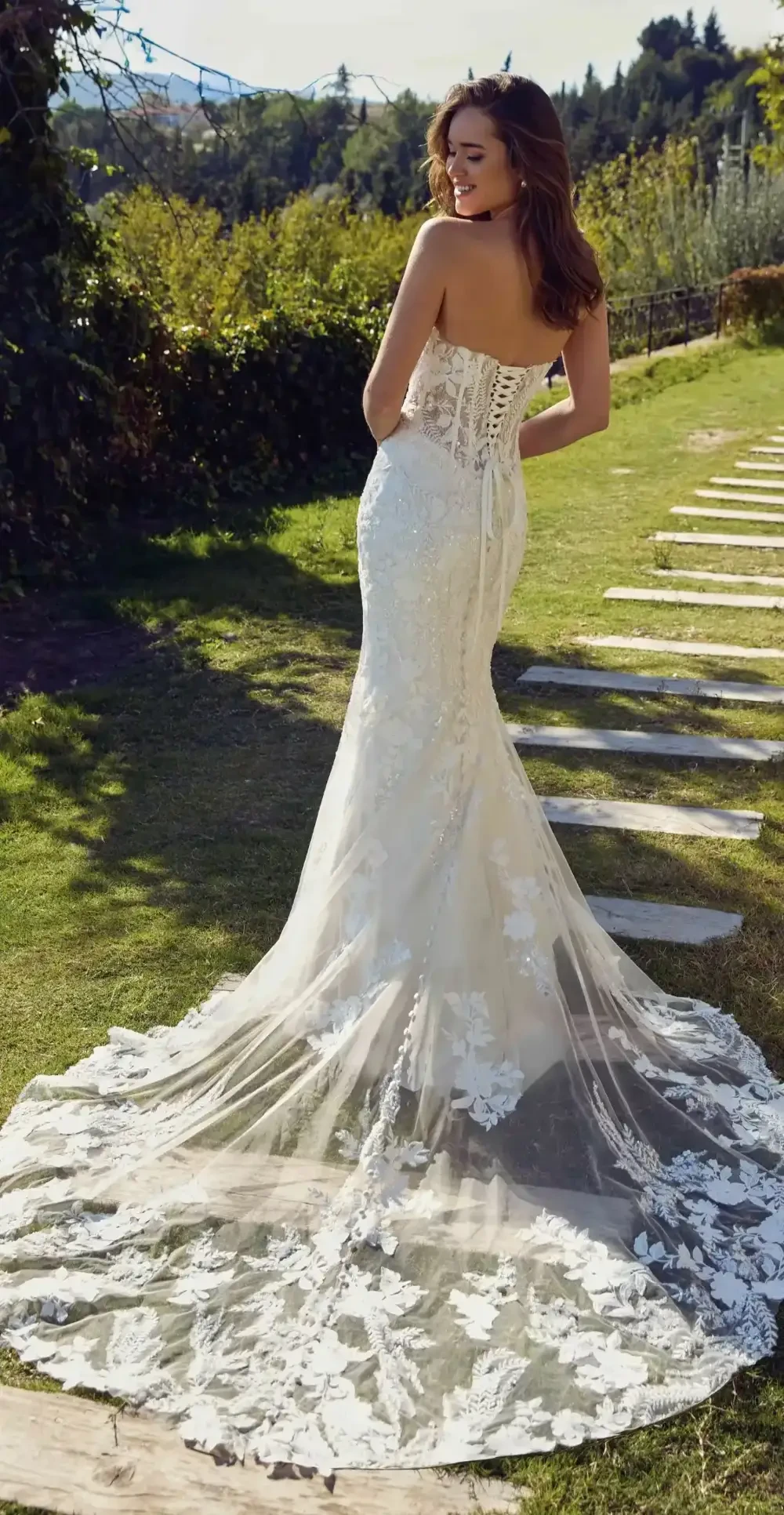 A woman is standing on a garden path, wearing a beautiful strapless wedding dress with intricate lace detailing, facing away from the camera. The background features greenery and a scenic landscape.