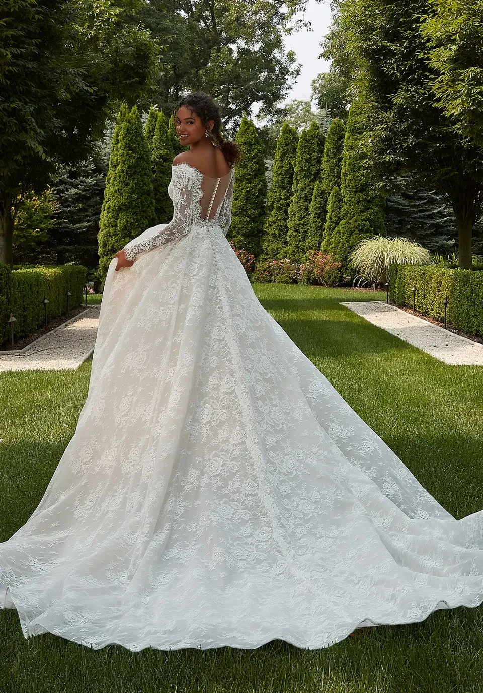 A woman in a lace off-shoulder wedding dress smiles warmly, standing in a lush garden. Sunlight filters through trees, creating a joyful, serene atmosphere.